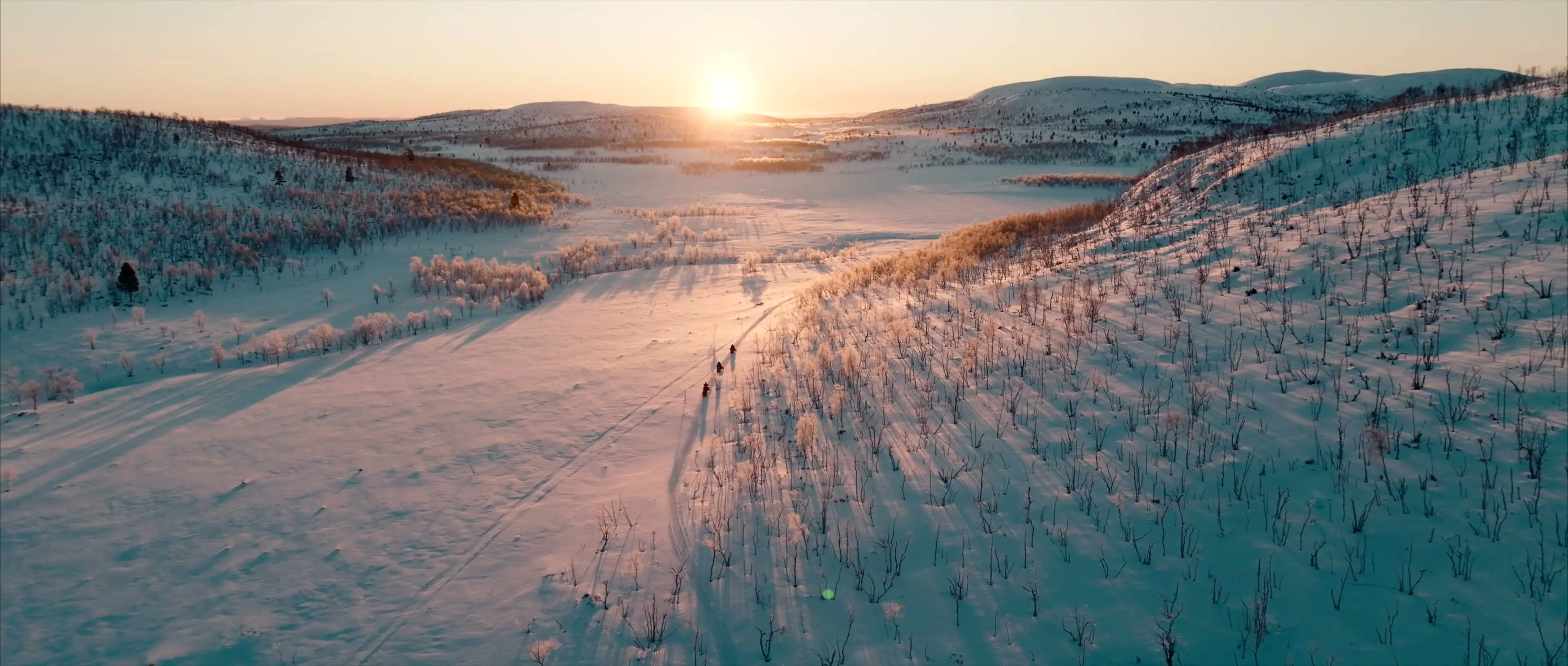 Three snowmobiles cross a wide, snowy valley towards the low winter sun