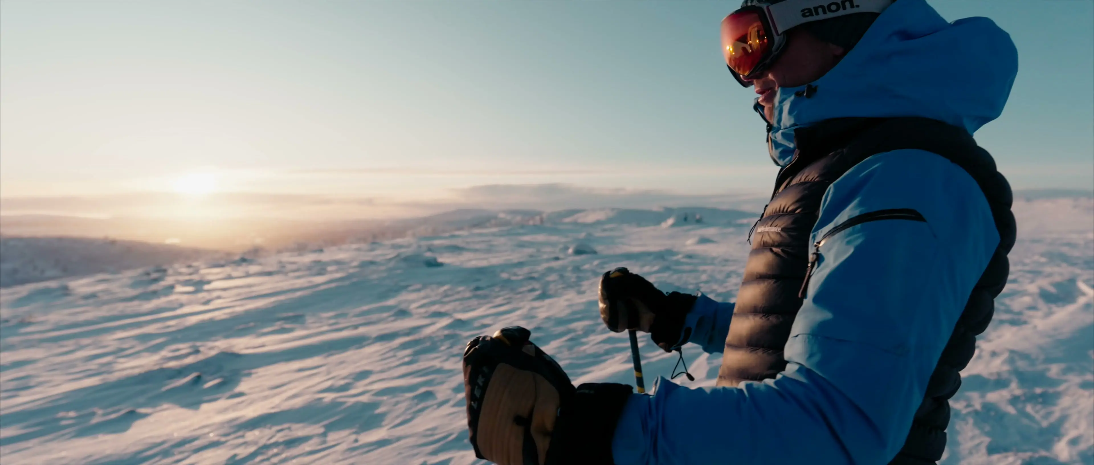 A skier in goggles resting on his poles on a snowy fell, the low winter sun on the horizon
