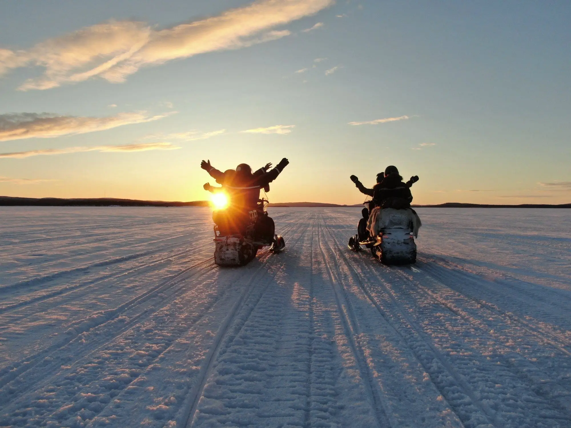 Two riders with their arms raised on snowmobiles on a frozen lake at sunset